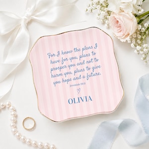 May include: A square ceramic trinket dish with wavy edges and a gold-toned rim. The surface features light pink vertical stripes and blue text that reads: For I know the plans I have for you, plans to prosper you and not to harm you, plans to give you hope and a future. Jeremiah 29:11, a small heart icon, and the name OLIVIA.