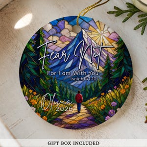 May include: A round ceramic ornament featuring a stained-glass style illustration of a person walking on a path toward a mountain. The text reads Fear Not For I am With You Isaiah 41:10, Olivia, and 2026. It includes a gold hanging string.