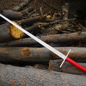 May include: A long, silver sword with a red handle and a silver crossguard. The sword is resting on a pile of logs. The blade is long and straight, and the handle is wrapped in red material.