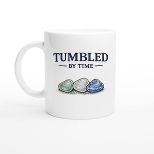 Könnte beinhalten: Ein weißer Keramikbecher mit dem dunkelblauen Schriftzug TUMBLED BY TIME über einer Illustration von drei glatten, abgerundeten Steinen in Grün-, Weiß- und Blautönen, die auf einem kleinen Haufen winziger Kieselsteine am Fuß des Motivs liegen.