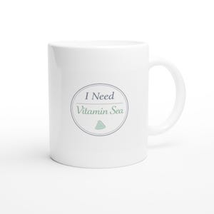 May include: A white ceramic mug featuring a circular design with a thin dark outline. Inside the circle, the text reads I Need Vitamin Sea in a dark blue and light green font, accompanied by a small light green triangle icon at the bottom.