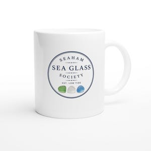 May include: A white ceramic mug featuring a circular navy blue logo that reads Seaham Sea Glass Society Est. Low Tide. Below the text, there are three small, smooth, rounded pieces of sea glass in shades of green, white, and blue.