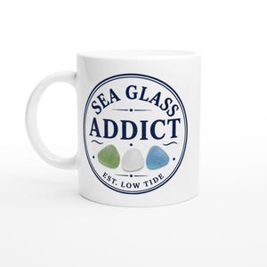 May include: A white ceramic mug featuring a circular navy blue graphic with the text SEA GLASS ADDICT and EST. LOW TIDE. The design includes three small, smooth, rounded sea glass pieces in shades of light green, white, and light blue.