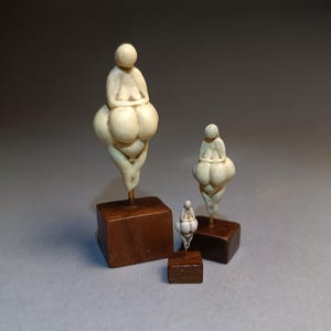 May include: Three ivory-coloured sculptures of stylised female figures, each mounted on a small, dark wooden base. The figures have exaggerated forms and are arranged in a row, varying in size.