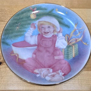 May include: Decorative plate featuring a smiling child in pink overalls and a white hat. The plate has a gold rim and a Christmas theme, with a decorated tree, a gift, and a blue background.
