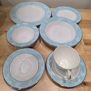 Peut inclure: Un ensemble de vaisselle blanche et bleu clair, comprenant des assiettes, des bols et une tasse à thé avec soucoupe. Les bords des plats ont une bordure bleu clair avec un motif de feuilles et une bordure argentée. Les plats sont disposés sur une surface en bois.