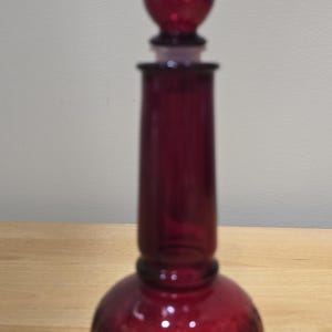 Op de afbeelding: Een dieprode glazen karaf met een hoge, slanke hals en een ronde basis met een gestructureerd ruitpatroon. De karaf is voorzien van een bijpassende, puntige en gestructureerde glazen stop die in de opening aan de bovenzijde past.