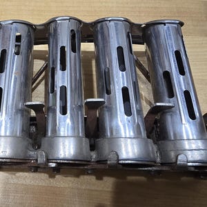 May include: A silver-coloured metal coin dispenser. It has four cylindrical compartments with rectangular slots and levers. The dispenser is mounted on a metal base and rests on a wooden surface.