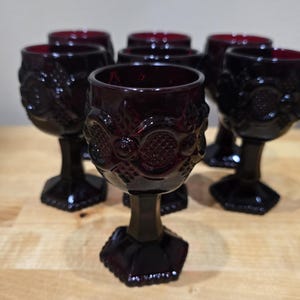 May include: A set of six deep red glass goblets featuring a textured pattern of raised circles and diamond cross-hatching. Each goblet has a thick, faceted hexagonal stem and a matching hexagonal base, crafted from translucent dark red glass.
