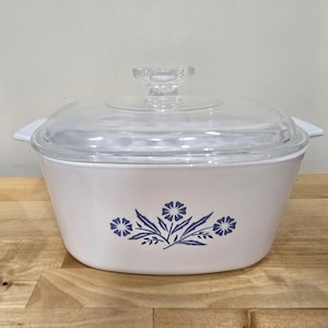 Könnte beinhalten: Weiße Auflaufform mit Glasdeckel und blauem Blumendesign. Das Design zeigt drei blaue Blumen mit grünen Blättern.