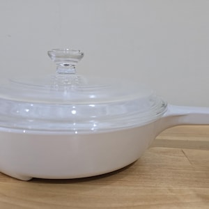 May include: A white saucepan with a clear glass lid and handle. The pan is on a wooden surface. The lid has a clear glass knob. The handle extends from the side of the pan. The pan is a kitchenware item.