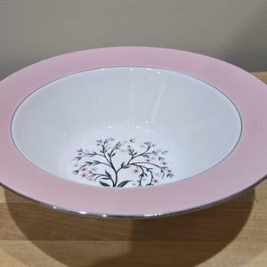 May include: A round, white ceramic bowl with a pink rim and a silver edge. The bowl has a floral design in the centre with a black tree and small pink flowers. The bowl is set on a wooden surface.