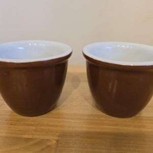 May include: Two brown ceramic ramekins with white interiors. The ramekins are sitting on a wooden surface.