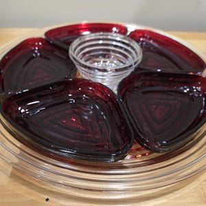 Può includere: Un vassoio di servizio in vetro trasparente con manici, contenente una piccola ciotola di vetro trasparente e quattro piatti triangolari in vetro rosso scuro. I piatti sono disposti attorno alla ciotola. Ideale per gli antipasti.