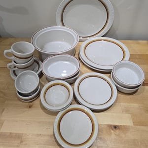 Op de afbeelding: Een verzameling crèmekleurig keramisch servies met bruine en oranje randen. De set bevat borden, kommen, kopjes en een ovenschaal. De borden variëren in grootte en de kopjes zijn opgestapeld.