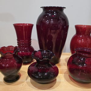以下が含まれることがあります： さまざまな形とサイズの6つのルビーレッドガラス花瓶のコレクション。花瓶は光沢のある仕上がりで、明るい色の木製の表面に配置されています。一部の花瓶にはフリルのリムがあります。