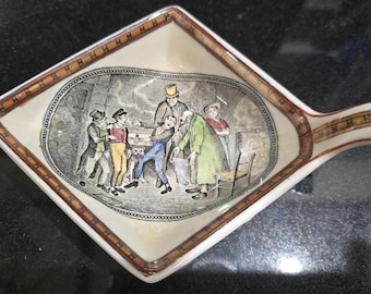 Może przedstawiać: Ceramiczne naczynie w kształcie rombu z uchwytem, z centralną ilustracją przedstawiającą pięć postaci w historycznych strojach oraz psa. Brzeg ozdobiony jest brązowo-białym wzorem w kratkę na kremowym tle.