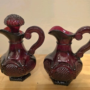 Könnte beinhalten: Zwei passende, tiefrote Glas-Ölkännchen mit verzierten Griffen und strukturierten Details. Eines hat einen dekorativen Verschluss. Die Kännchen haben einen Ausguss zum Ausgießen und stehen auf einem quadratischen Sockel. Das Design ist Vintage.