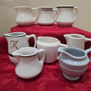 Puede incluir: Una colección de jarras de crema de cerámica blanca con varios diseños. Algunas jarras tienen adornos negros o dorados, y una tiene una "K" dorada. Una jarra es de color azul claro.