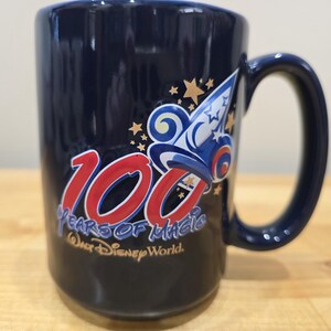 May include: A navy blue ceramic mug with a large handle. The mug features the text "100 Years of Magic" in red and white, along with a graphic of a wizard's hat and stars. The words "Walt Disney World" are also visible.