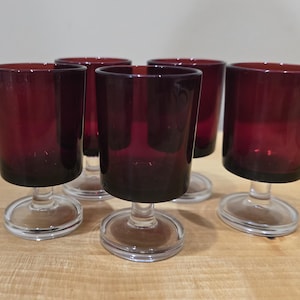 May include: A set of five small drinking glasses featuring deep burgundy red bowls with smooth, cylindrical sides. Each glass is supported by a short, clear glass stem and a circular, transparent base, creating a uniform look for the collection.