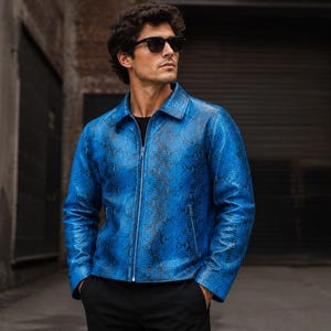 May include: A vibrant blue jacket featuring a detailed snakeskin pattern in black. This long-sleeved garment includes a classic collar, a full-length silver-toned front zipper, and two side pockets with matching silver zippers, worn over a black shirt.