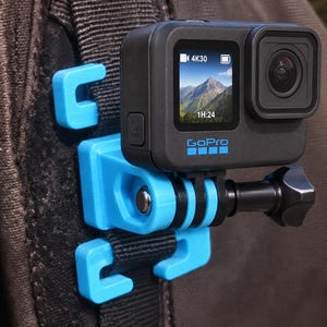 May include: A black GoPro action camera mounted on a bright blue plastic clip. The camera screen displays a mountain landscape with 4K30 and 1H:24 text. The blue mount features a textured surface and is attached to a dark brown fabric strap.