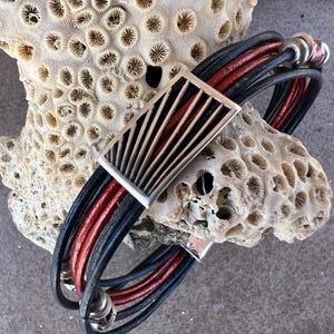 May include: A multi-strand bracelet featuring black and red leather cords. The bracelet has a silver-tone rectangular clasp with a fan-like design. The bracelet is displayed on a piece of coral.