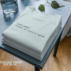 May include: A stack of white t-shirts with the text "[USER_ERROR] Problem not found in system." printed in black. The shirts are neatly folded on a light blue table. The words "HEATHER PRISM ICE BLUE" are at the bottom.