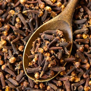 May include: A wooden spoon filled with dried whole cloves, resting on a dense bed of additional dried whole cloves. The cloves are dark brown with lighter tan, rounded, four-pointed flower bud tops, showcasing a textured, organic appearance.