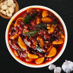 Könnte beinhalten: Eine weiße Schüssel gefüllt mit dunkelrotem, scharfem Knoblauch-Pickle, bestehend aus ganzen Knoblauchzehen, einer dunkelroten Chilisauce und grünen Curryblättern. Daneben steht eine kleine Holzschale mit geschälten Knoblauchzehen und Knoblauchknollen.