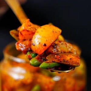 Puede incluir: Una cuchara de metal sostiene una porción de encurtido indio picante, con trozos de mango amarillo, pequeños chiles verdes y especias oscuras cubiertas con un aceite rojo vibrante, extraído de un frasco de vidrio lleno de la misma mezcla.