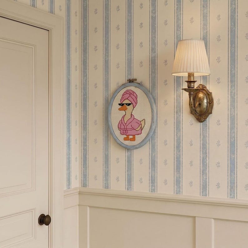 May include: An oval embroidery hoop containing a cross-stitch pattern of a duck wearing a pink bathrobe, a matching pink towel wrapped around its head, and black sunglasses. The duck has orange feet and a yellow beak, set against a white fabric background.
