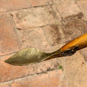 May include: A handcrafted wooden spoon with a brass-coloured scoop. The handle is a light brown colour, and the scoop has a leaf-like shape. The spoon is held in a hand against a background of brick pavers.
