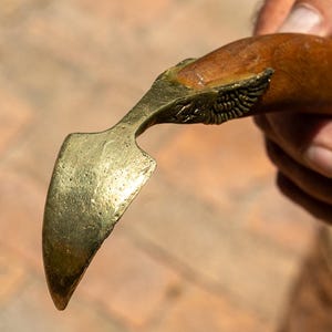May include: A vintage brass and wood knife with a curved blade. The handle is a dark brown colour, and the blade is a shiny gold. The knife is held in a hand, with a blurred background of a brick surface and a terracotta pot with plants.