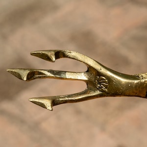 May include: A close-up of a trident head made of gold-coloured metal. The trident has three prongs and is attached to a wooden handle. The handle is a dark brown colour. The trident is a decorative item.