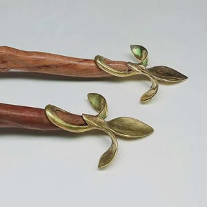 May include: Two wooden bottle stoppers with a fish carving on the end. Each stopper has a brass-coloured, leaf-shaped design. The wood is a medium brown colour, and the brass has a slight green tint.