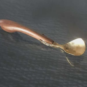 May include: A spoon with a wooden handle and a brass bowl. The handle is curved and made of light brown wood. The bowl is a shiny gold colour. The spoon is likely a serving utensil.