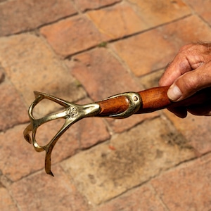May include: A brass and wood tool with a four-pronged head and a wooden handle. The brass head has an ornate design. The handle is a dark wood colour. The tool is held in a hand against a brick background.