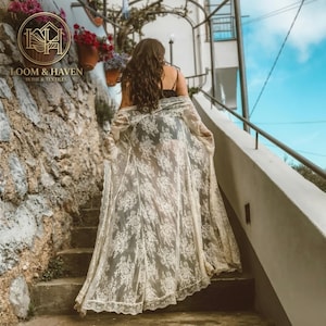 May include: A floor-length, sheer white lace duster featuring an intricate floral pattern, long sleeves, and an open front design. The garment is worn over a dark base layer, showcasing a delicate scalloped hemline and lightweight fabric.