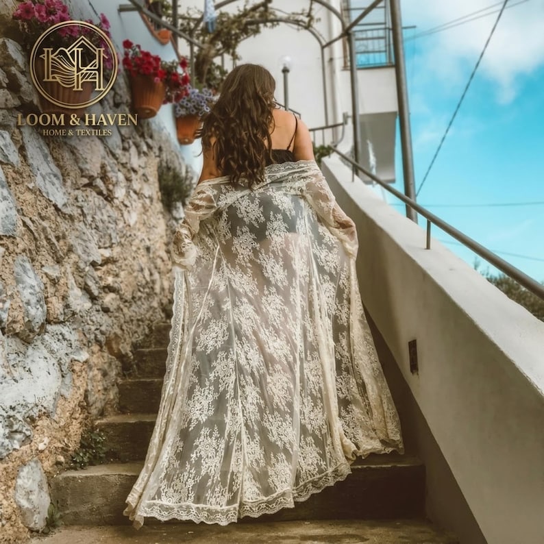 May include: A floor-length, sheer white lace duster featuring an intricate floral pattern, long sleeves, and an open front design. The garment is worn over a dark base layer, showcasing a delicate scalloped hemline and lightweight fabric.