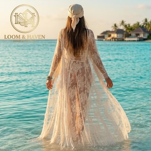 May include: A floor-length, sheer beige lace cover-up featuring long sleeves, a tie-waist closure, and intricate floral patterns. The garment is worn with a matching beige headscarf, showcasing a delicate, semi-transparent design.