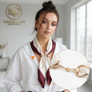 May include: A square silk scarf featuring a cream center and a deep burgundy border, secured at the neck with a polished gold-tone metal horsebit-style ring clasp. The scarf is worn over a crisp white collared shirt, with a circular inset detail.