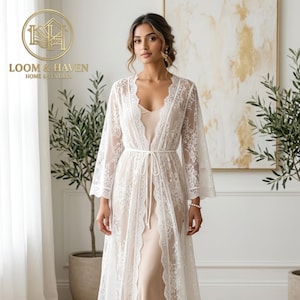 May include: A floor-length robe featuring white floral lace overlay with wide sleeves and a thin waist tie, worn over a solid beige slip dress. The garment has a scalloped lace trim along the front opening and a V-neckline, creating a layered look.