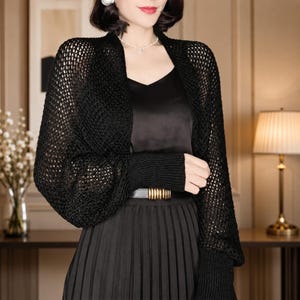 May include: A black open-front shrug featuring a loose, net-like crochet knit pattern, long sleeves with wide ribbed cuffs, and a cropped silhouette. The garment is worn over a black sleeveless top and a black pleated skirt with a gold-toned belt.
