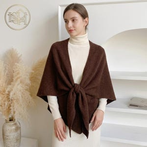 May include: A brown shawl tied in the front, worn over a white turtleneck and dress. The shawl is a solid color and appears to be made of a soft material. The background includes a vase with tall, dried plants and a white wall.