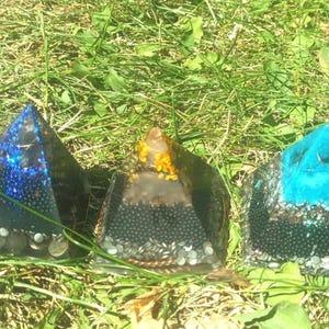 Op de afbeelding: Vijf piramidevormige objecten van hars, elk met ingebedde metaalsnippers, kleine steentjes en glitters. De kleuren zijn rood, donkerblauw, amber en twee tinten helder turkoois, allemaal met een gelaagde interne samenstelling.