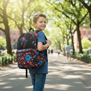 K&ouml;nnte beinhalten: Ein schwarzer Rucksack mit Spider-Man. Der Rucksack hat ein gro&szlig;es Bild von Spider-Man auf der Vorderseite und ein Muster aus Spider-Man-Logos und -Symbolen. Der Rucksack wird von einer Person auf einem von B&auml;umen ges&auml;umten Weg getragen.