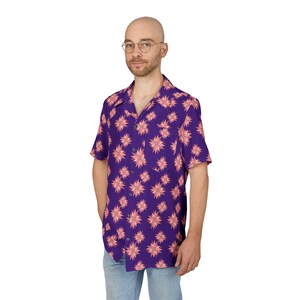 May include: A short-sleeved purple shirt with a repeating pattern of pink starburst-like designs. The shirt has a classic collar and button-down front. The model is wearing blue jeans.