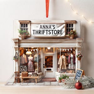May include: A flat, acrylic ornament shaped like a storefront with a sign reading ANNA'S THRIFTSTORE. The miniature shop features display windows with mannequins wearing clothing, a central door, potted plants, and signs that say SPECIAL DEALS.